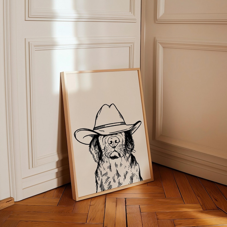 May include: Black and white line art print of a dog wearing a cowboy hat. The print is framed in a light wood frame and is leaning against a white wall.