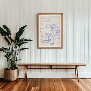 May include: A framed blue and white art print depicts a person relaxing with a cocktail on a beach. The artwork is in a light wood frame and hangs on a white paneled wall. A wooden bench with a woven top sits below the artwork.