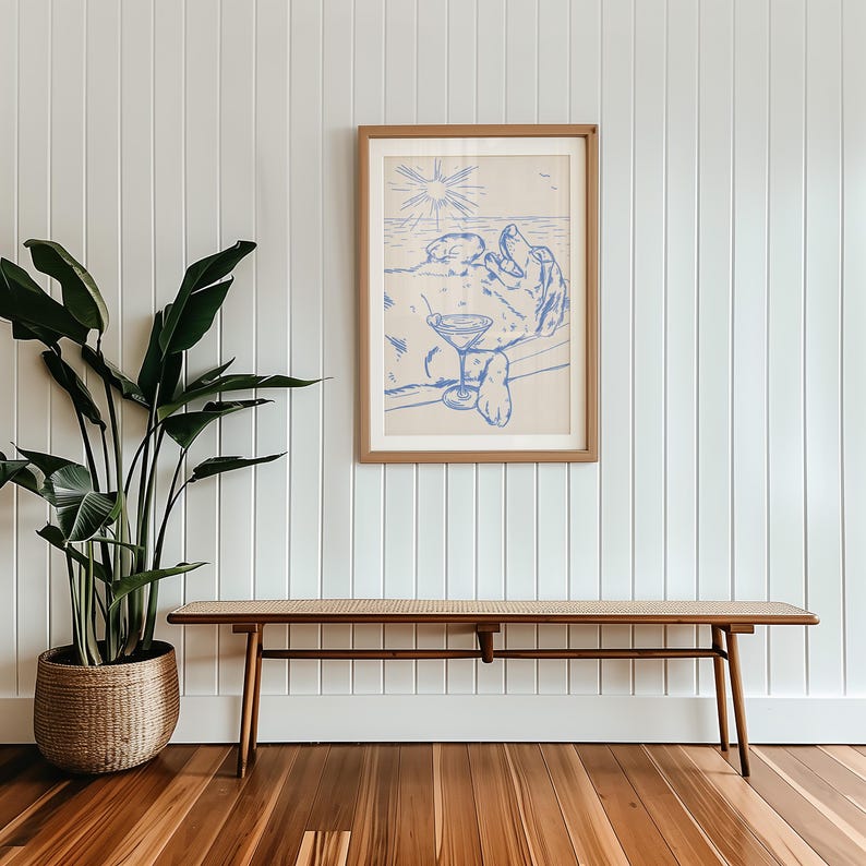 May include: A framed blue and white art print depicts a person relaxing with a cocktail on a beach. The artwork is in a light wood frame and hangs on a white paneled wall. A wooden bench with a woven top sits below the artwork.