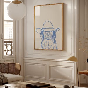 May include: A framed print featuring a blue illustration of a dog wearing a cowboy hat. The artwork has a light beige background and a gold frame. The dog's face is detailed with blue lines, creating a whimsical and rustic aesthetic. The print is displayed in a well-lit room.