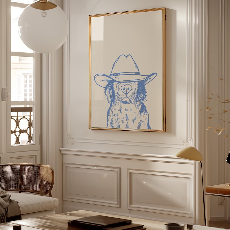 May include: A framed print featuring a blue illustration of a dog wearing a cowboy hat. The artwork has a light beige background and a gold frame. The dog's face is detailed with blue lines, creating a whimsical and rustic aesthetic. The print is displayed in a well-lit room.