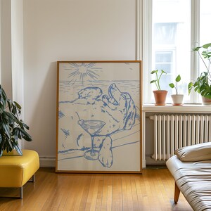 May include: A framed art print featuring a blue line drawing of a dog relaxing with a martini on a beach. The artwork includes a sun, ocean, and a dog lounging on a beach chair. The frame is made of wood.