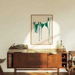 May include: A framed illustration of teal-colored bikini tops hanging on a clothesline against a beige background. The illustration is displayed on a wooden cabinet with a record player on top.