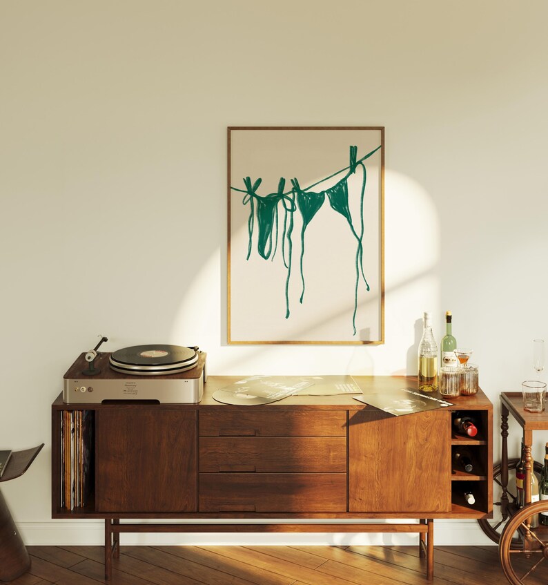 May include: A framed illustration of teal-colored bikini tops hanging on a clothesline against a beige background. The illustration is displayed on a wooden cabinet with a record player on top.
