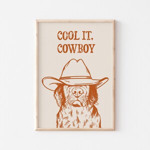Op de afbeelding: Een printbaar wanddecoratiedesign met een hond die een cowboyhoed draagt met de tekst "COOL IT, COWBOY" in oranje op een beige achtergrond.