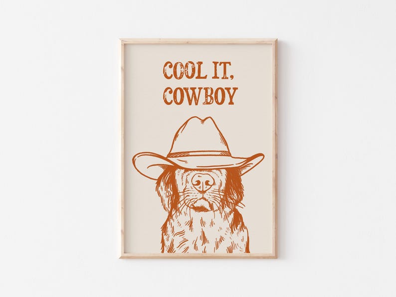 Op de afbeelding: Een printbaar wanddecoratiedesign met een hond die een cowboyhoed draagt met de tekst "COOL IT, COWBOY" in oranje op een beige achtergrond.