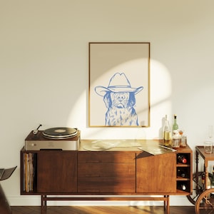 May include: A framed print featuring a blue line drawing of a dog wearing a cowboy hat. The artwork is displayed above a mid-century modern wooden cabinet with a record player and vinyl records. The room has a warm, inviting atmosphere.