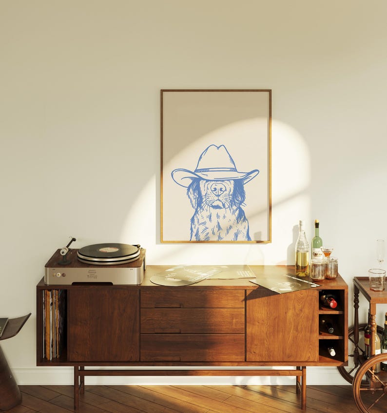 May include: A framed print featuring a blue line drawing of a dog wearing a cowboy hat. The artwork is displayed above a mid-century modern wooden cabinet with a record player and vinyl records. The room has a warm, inviting atmosphere.
