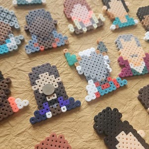 May include: A selection of colourful pixel art magnets, depicting different figures and designs. The magnets are crafted from small, fused plastic beads in shades of brown, blue, white, and other colours. They are arranged on a textured, brown surface.