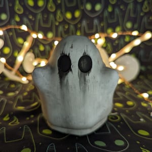 Op de afbeelding: Een klein, wit spookfiguur met zwarte ogen en donkere strepen. Het spook staat tegen een donkere achtergrond met een herhalend patroon van groene en zwarte katachtige figuren en een slinger van warmwitte lampjes.