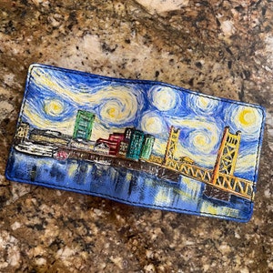 May include: A blue leather wallet with a painted image of the Portland skyline, including the iconic St. Johns Bridge, in a style reminiscent of Van Gogh's Starry Night. The wallet has a stitched border and the artist's signature in the bottom right corner.