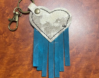 May include: A white leather heart-shaped keychain with a blue fringe. The keychain has a silver metal ring and a brown leather strap.