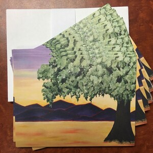 May include: A stack of blank greeting cards with a sunset landscape design. The cards feature a green tree with a thick trunk, purple mountains, and a yellow sky.