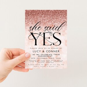 a hand holding a pink and gold invitation card with text and a glittery background.