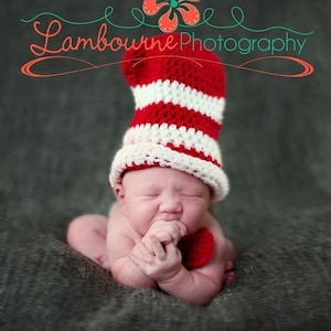 May include: A newborn baby wearing a red and white striped crocheted hat with a red crocheted bow tie. The baby is sitting on a gray textured surface.