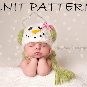 May include: A white knitted snowman hat with green ear flaps and a pink bow. The hat has black eyes, a yellow nose, and a black mouth. The hat is being worn by a baby.