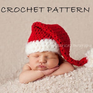 May include: A newborn baby wearing a red and white crocheted Santa hat with a white pom-pom on the end. The baby is sleeping with their arms crossed in front of them.