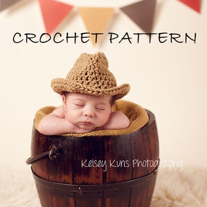 May include: A newborn baby wearing a crocheted cowboy hat, resting in a wooden barrel. The barrel is dark brown with metal bands. The words "CROCHET PATTERN" are above the baby. A banner of triangles hangs in the background.