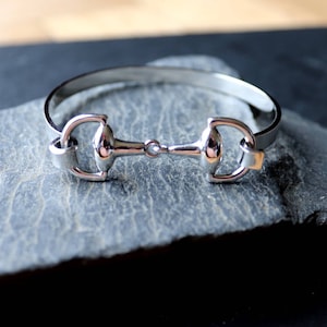 May include: A silver-toned bangle bracelet featuring a horse bit design. The bracelet is made of metal and has a polished finish.