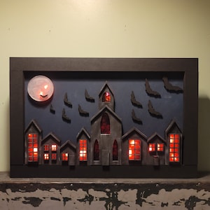 May include: A black framed shadow box with a Halloween village scene. The scene includes a church, houses, a moon, and bats. The windows of the buildings are lit with red lights.