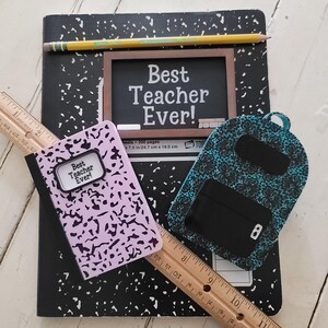 May include: A pink and black composition notebook with the words "Best Teacher Ever!" on the cover. A blue and black backpack keychain with a black and white floral pattern. A black and white composition notebook with the words "Best Teacher Ever!" on a black chalkboard.