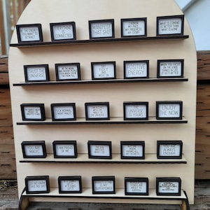 Könnte beinhalten: Ein Holzregal mit mehreren kleinen, gerahmten Schildern. Jedes Schild hat einen schwarzen Rahmen und weißen Text mit motivierenden Sätzen wie „Manifestiere diesen Mist“ und „Vertraue dem Universum“.