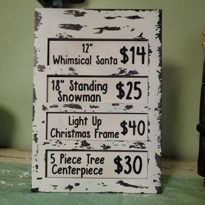 May include: A white wooden sign with black text listing prices for various Christmas decorations. The sign includes a 12 inch whimsical Santa for $14, an 18 inch standing snowman for $25, a light up Christmas frame for $40, and a 5 piece tree centerpiece for $30.