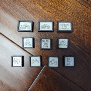 May include: A selection of small, square signs with black frames and white text. The signs display book-related quotes and phrases, including "Life is better with books & dogs" and "Just one more chapter."