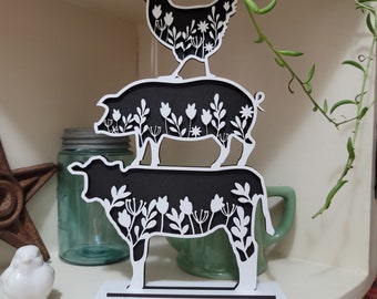 May include: A black and white wooden cutout of a chicken, pig, and cow stacked on top of each other. Each animal has a floral design.
