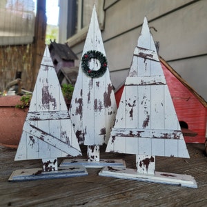 Op de afbeelding: Drie witgekalkte houten kerstboom met verweerde verf en een rustieke uitstraling. De middelste boom heeft een groene krans met rode bessen.