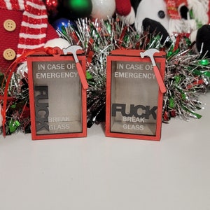 Puede incluir: Dos cajas de madera con marco rojo con el texto "IN CASE OF EMERGENCY" y "FUCK BREAK GLASS". Cada caja tiene un pequeño martillo adjunto. Las cajas están decoradas con cinta roja y oropel.