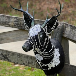 Puede incluir: Una cabeza de ciervo negra con cuernos, adornada con encaje blanco y cadenas plateadas. Los ojos del ciervo est&aacute;n pintados de blanco, y las cadenas est&aacute;n decoradas con peque&ntilde;as perlas.
