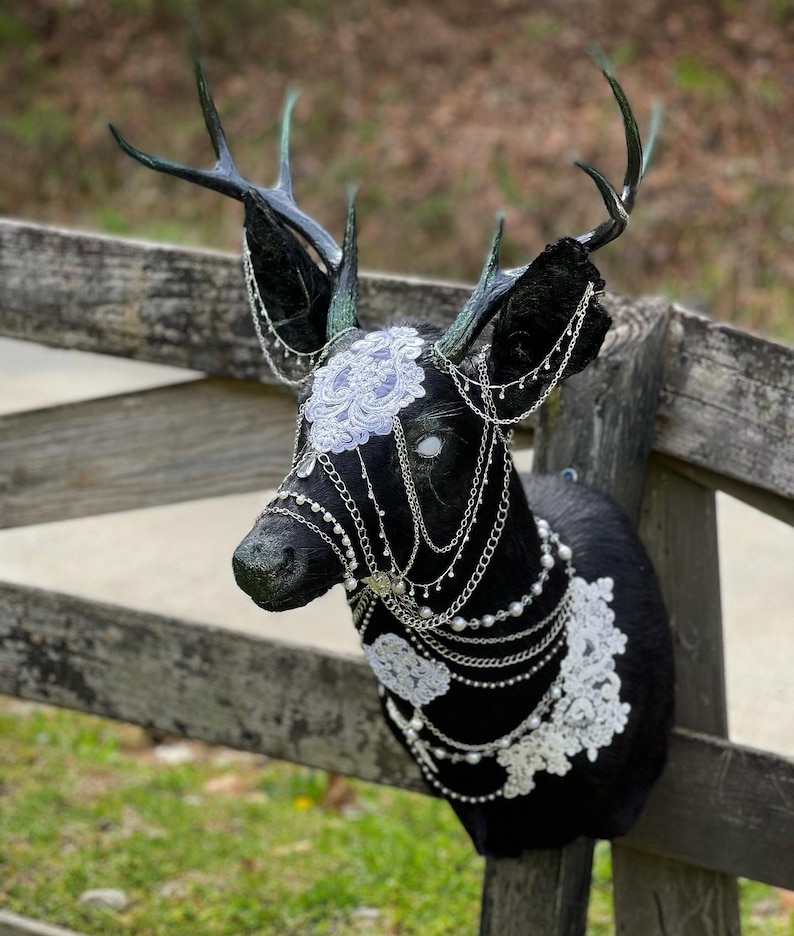 Puede incluir: Una cabeza de ciervo negra con cuernos, adornada con encaje blanco y cadenas plateadas. Los ojos del ciervo est&aacute;n pintados de blanco, y las cadenas est&aacute;n decoradas con peque&ntilde;as perlas.