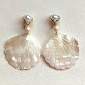 May include: A pair of clip-on earrings with a seashell design. Each earring features a pearlescent, fan-shaped shell pendant, a small pearl, and a larger pearl at the top. The earrings have a gold-coloured metal clip-on backing.