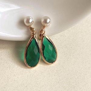 May include: A pair of elegant drop earrings. Each earring features a pear-shaped, faceted emerald green gemstone set in a gold-tone frame. A small, round, white pearl tops each earring. The earrings are displayed on a white surface.