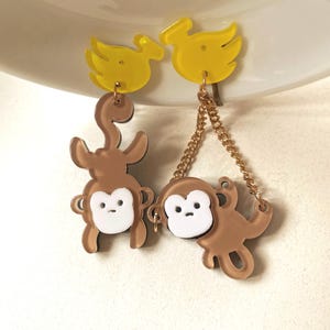 May include: A pair of whimsical earrings featuring yellow bird-shaped tops and brown monkey charms. The monkeys have white faces and are suspended from the birds by gold-colored chains. The earrings are designed with a playful, cartoonish aesthetic.