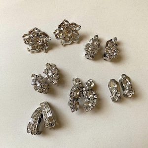 May include: A collection of five pairs of silver-tone clip-on earrings featuring clear rhinestones in various shapes, including floral, marquise, and baguette cuts. The set includes diverse designs with faceted crystals set in metal frames.