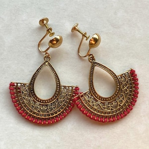 May include: A pair of gold-toned clip-on earrings with a teardrop and fan design. The earrings feature intricate filigree patterns and are accented with a row of small, bright red beads along the bottom edge. The earrings are designed for non-pierced ears.