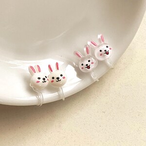 May include: Clip-on earrings shaped like white bunnies with pink details. The earrings have a cartoon design with black eyes and a smiling mouth. The earrings are presented on a white plate.