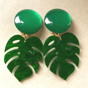 May include: Clip-on earrings with a large, round, glossy green top and a leaf-shaped pendant. The pendant is a vibrant green, resembling a Monstera leaf. The earrings have a gold-coloured clip.