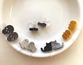 以下が含まれることがあります： ハロウィーンをテーマにしたイヤリングのコレクション。スカル、ゴースト、猫、カボチャのデザインが含まれています。イヤリングは黒、シルバーグリッター、ゴールドグリッターです。イヤリングは白いお皿に飾られています。