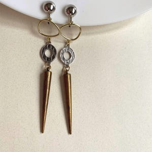 May include: Dangling earrings featuring silver ball studs, gold teardrop shapes, silver circular designs, and gold cone pendants. The earrings are displayed on a white surface.