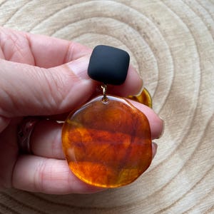 May include: A pair of statement earrings featuring a large, round, amber-coloured resin piece and a small, square, black resin piece.