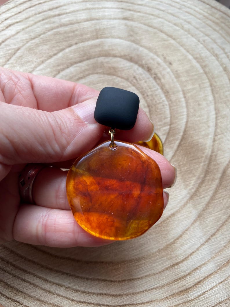 May include: A pair of statement earrings featuring a large, round, amber-coloured resin piece and a small, square, black resin piece.