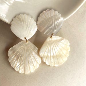May include: A pair of drop earrings featuring two stacked white iridescent shell-shaped components. Each earring consists of a smaller top shell and a larger bottom shell connected by a small gold-toned metal jump ring, creating a layered design.
