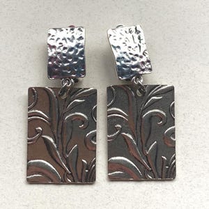 May include: A pair of silver-coloured earrings. Each earring has a rectangular, textured top piece and a larger, rectangular pendant with an embossed floral design. The earrings are connected by a small, circular link.