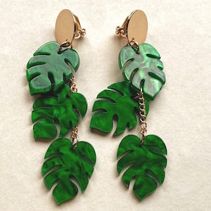 May include: A pair of clip-on earrings featuring a tropical leaf design. The earrings have gold-colored circular clips and chains, with multiple green, leaf-shaped pendants. The leaves have a marbled effect.