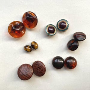 May include: A collection of assorted round buttons in various sizes and finishes. Included are translucent amber-coloured buttons, small tiger-eye patterned buttons, dark brown textured buttons with a snakeskin-like pattern, and dark purple buttons.