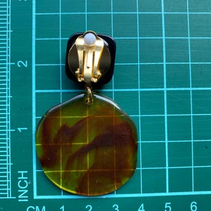 May include: A pair of clip-on earrings with a large, round, translucent green and brown resin design. The earrings have a gold-tone clip closure.