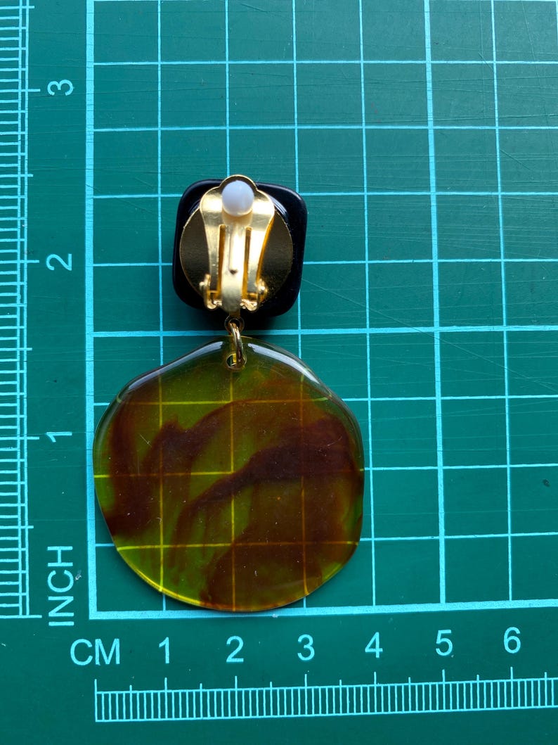 May include: A pair of clip-on earrings with a large, round, translucent green and brown resin design. The earrings have a gold-tone clip closure.